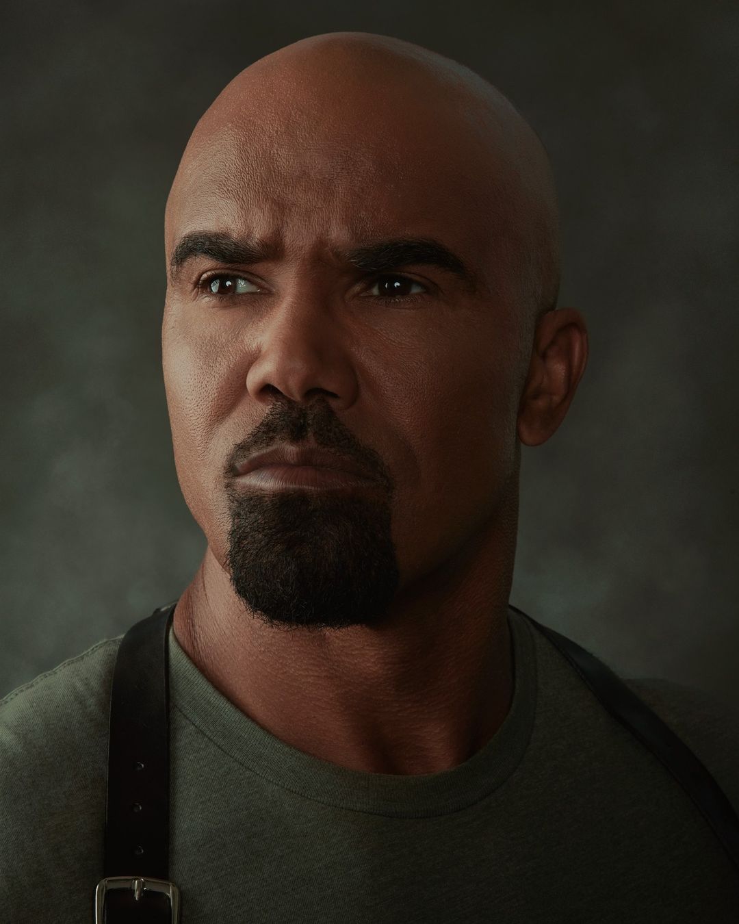 Close-up portrait of an adult male with a shaved head and goatee wearing a green shirt and black suspenders against a gray backdrop.